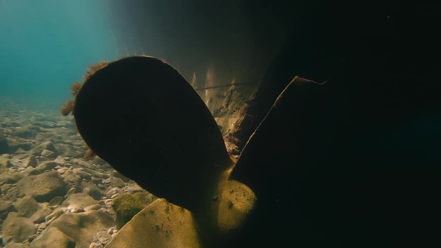 An eerie underwater view pans slowly past a massive ship's propeller and along its dark, algae-covered hull