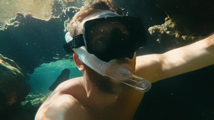 A man captures a slow-motion selfie video while snorkeling backwards through a sunlit underwater cave