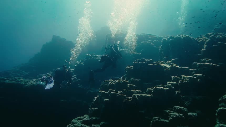A stunning cinemagraph of two scuba divers exploring a majestic underwater rock formation as streams of bubbles rise towards the sunlit surface