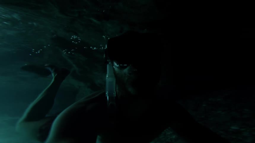 An adventurous man films a selfie video of himself snorkeling through a dark, narrow underwater sea cave