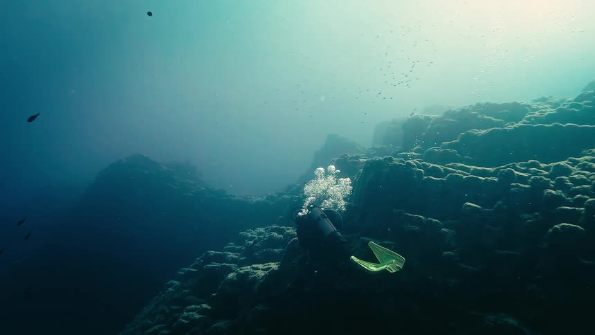 A slow-motion underwater view of a scuba diver picking up a discarded plastic bottle from a rocky ocean floor