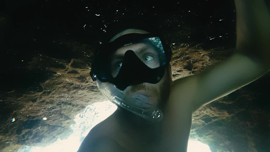 A man with a snorkel mask takes a selfie video from a low angle while floating inside a dark underwater sea cave