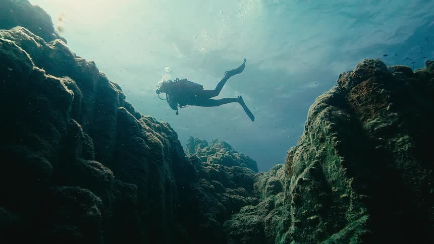 A silhouetted scuba diver explores a deep underwater canyon illuminated by sunlight from above