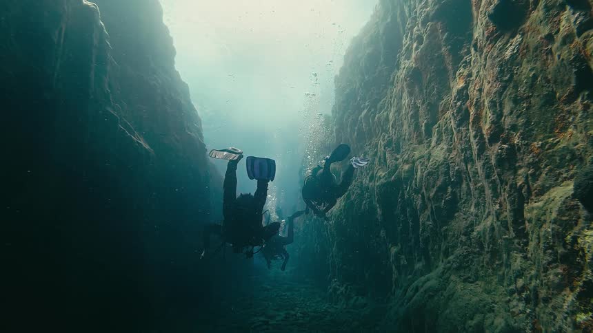 Scuba divers navigate a dark and mysterious underwater canyon as ethereal light streams from above