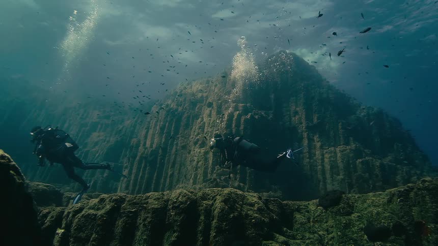 Two scuba divers in slow motion explore a majestic, ridged underwater mountain surrounded by schools of small fish