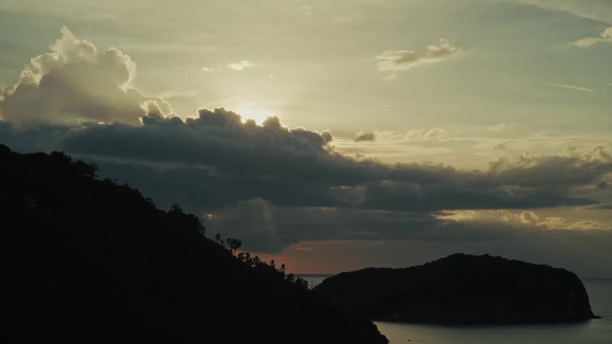 The sun sets behind dramatic dark clouds over a tranquil ocean, casting coastal hills and an island into silhouette