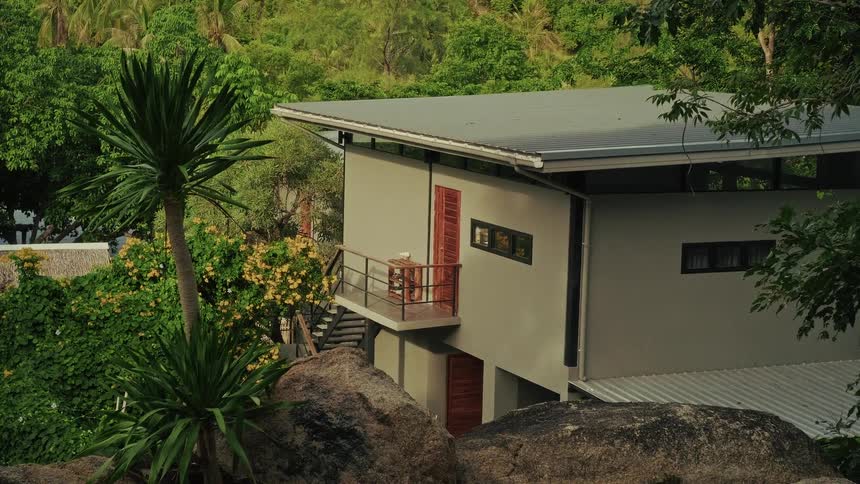 A serene, modern tropical villa with a balcony is nestled amongst lush green trees and large boulders