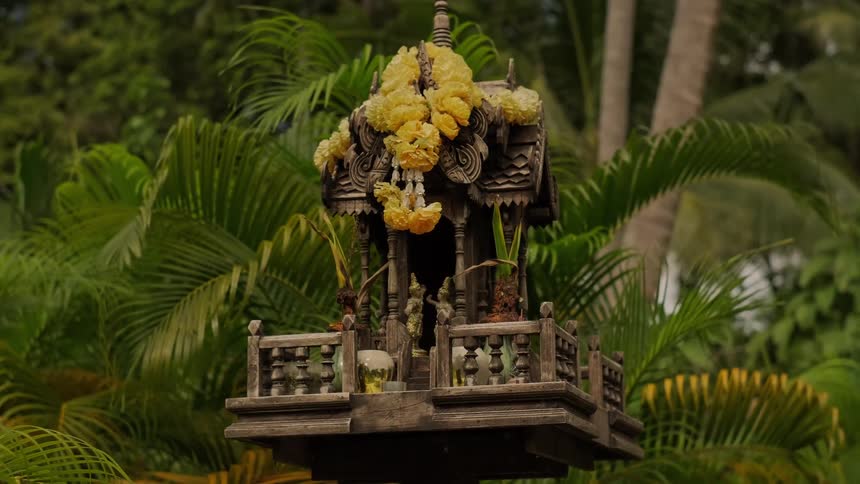 A traditional Thai wooden spirit house decorated with yellow flower garlands sits among lush tropical palm trees