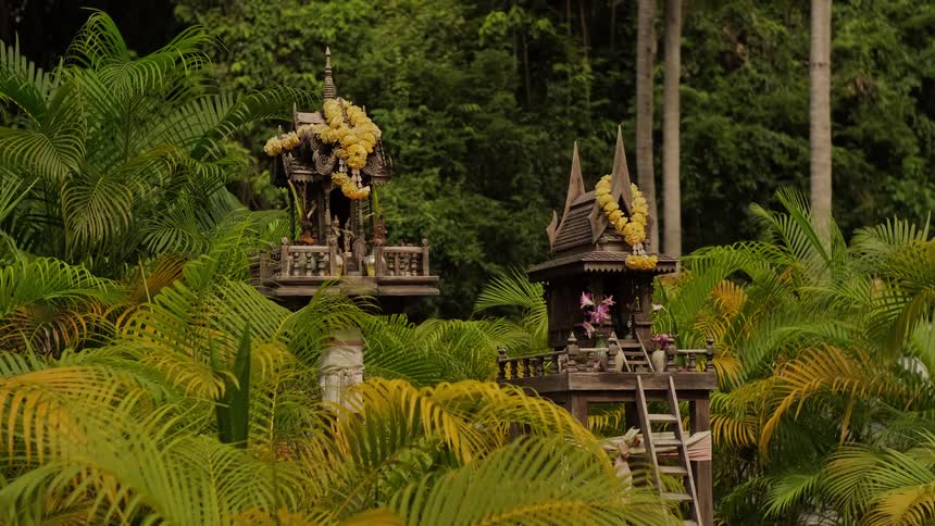 Two traditional Thai spirit houses adorned with yellow flower garlands sit peacefully among lush tropical palm trees in a jungle setting