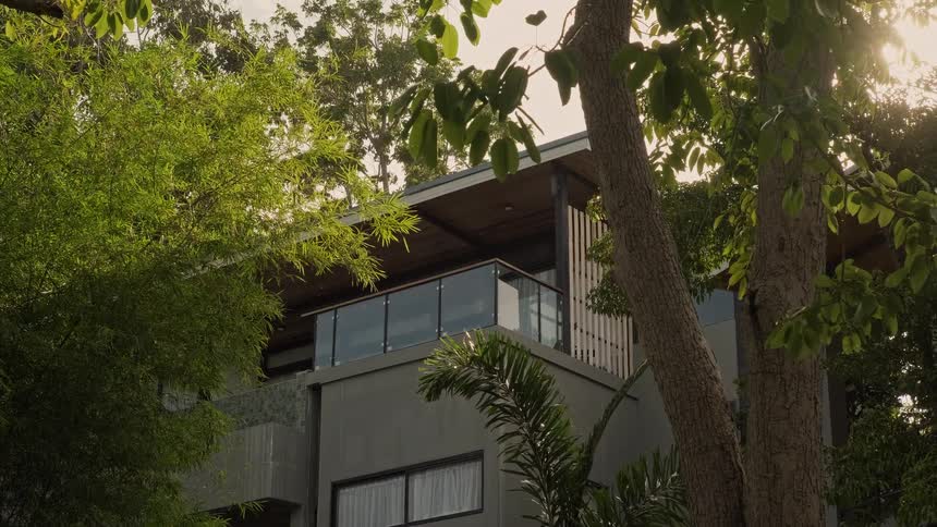 A modern luxury home with a glass balcony is seen through lush green trees as the sun shines brightly
