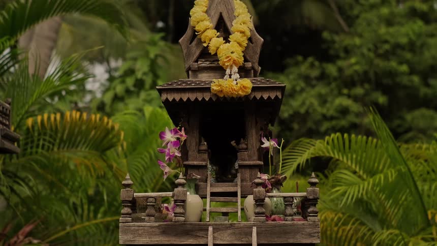 A traditional Thai wooden spirit house adorned with yellow marigold garlands and fresh orchids sits peacefully in a lush tropical garden