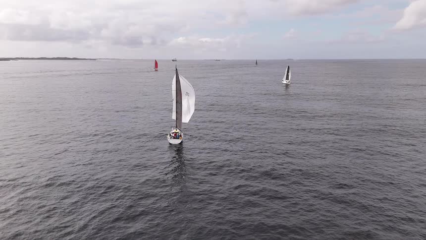 A stunning aerial shot captures a sailboat gliding across the dark ocean during a competitive regatta under a cloudy sky