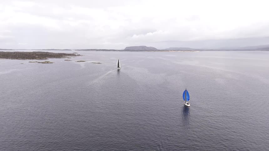 Static aerial shot of two sailboats with colorful spinnakers racing across a vast fjord on a cloudy day
