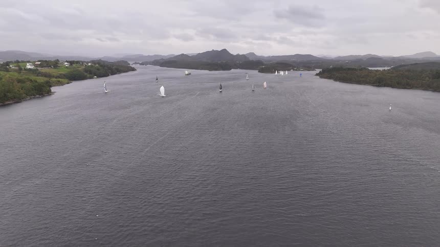 An aerial view shows multiple sailboats racing in a regatta on a wide fjord on a calm, overcast day