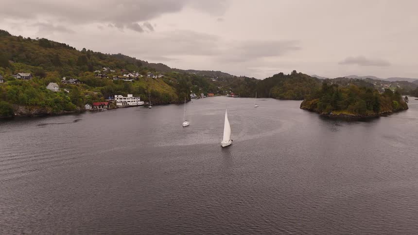 A serene aerial shot follows a sailboat as it navigates a tranquil fjord, surrounded by forested hills and a coastal town on an overcast day
