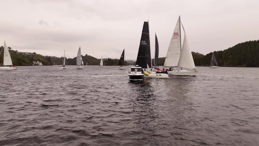 A fleet of sailboats and a motorboat navigate a choppy lake during a regatta on an overcast day