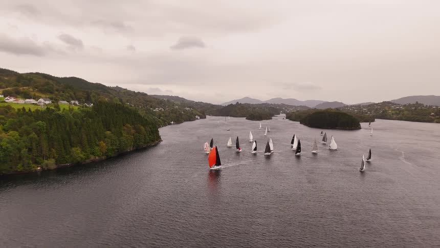 Aerial view of a sailboat regatta on a vast fjord, with one boat's vibrant orange spinnaker standing out on a cloudy day