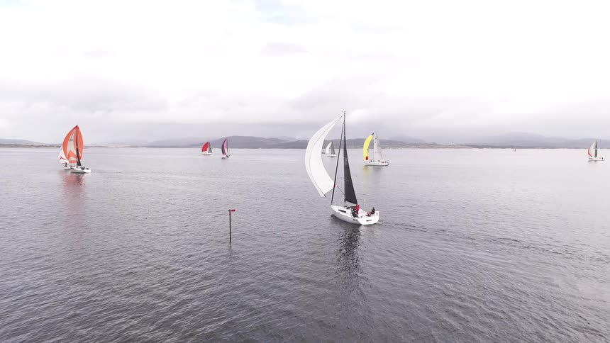 An aerial view follows a white sailboat as it competes with other yachts in a regatta on a calm, overcast day