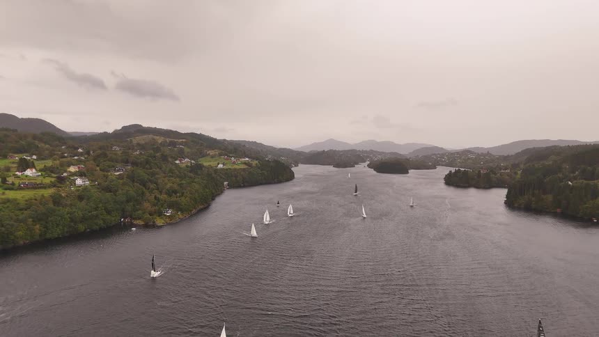 An aerial view captures a fleet of sailboats gliding across a tranquil lake during a regatta on a cloudy day