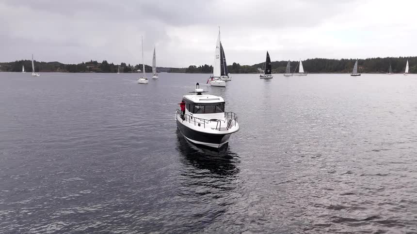 A black and white cabin cruiser floats on a gray sea as a fleet of sailboats pass in the background under an overcast sky