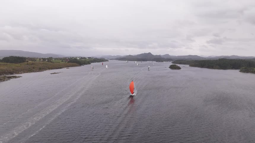 Aerial view of a sailboat regatta on a wide fjord on a cloudy day, with a striking orange spinnaker in the foreground