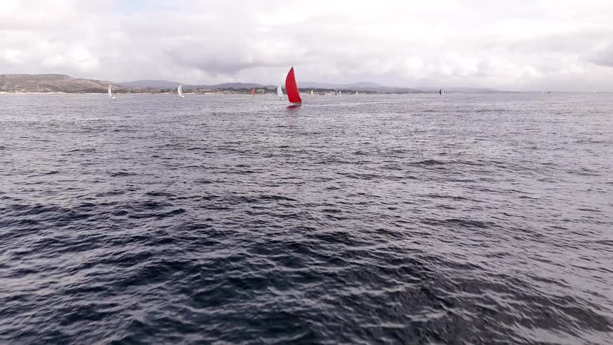 Flying low over the ocean, the camera tilts up to reveal a fleet of sailboats racing in a regatta under a cloudy sky