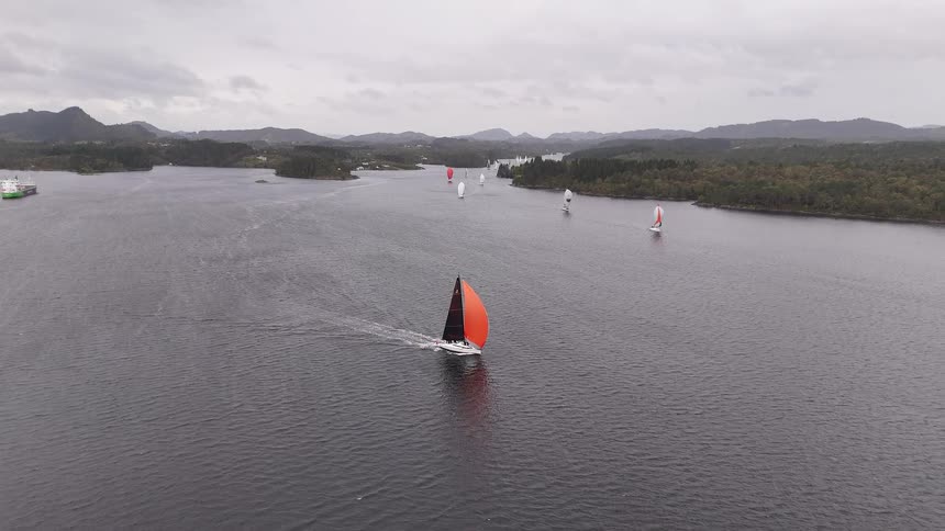 A sailboat with a vibrant orange spinnaker sail glides across the water, leading a fleet of boats during a regatta under an overcast sky