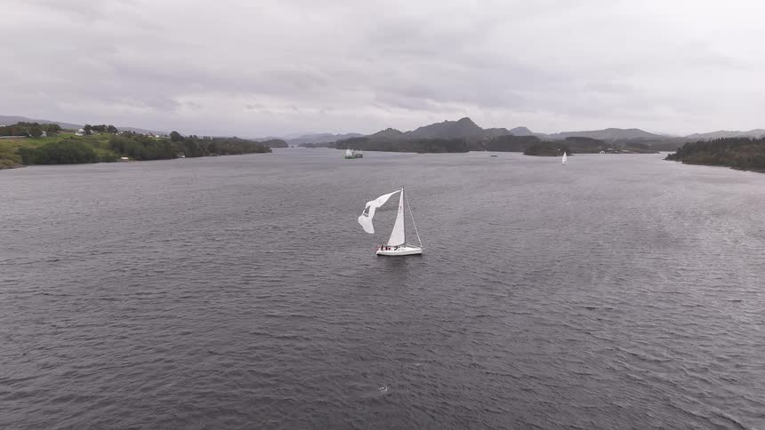 A majestic white sailboat gracefully navigates the dark, expansive waters of a fjord on a cloudy, overcast day