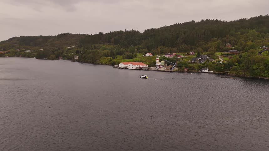 Aerial view of a small boat approaching an industrial coastal village at the base of a forested hill on a cloudy day