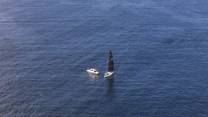 A high-angle view shows a sleek sailboat with a black sail gliding across the calm, open ocean, accompanied by a small motorboat