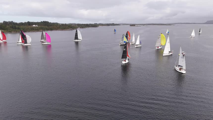 A static aerial shot shows a large fleet of sailboats with colorful spinnakers competing in a regatta on a calm, overcast day