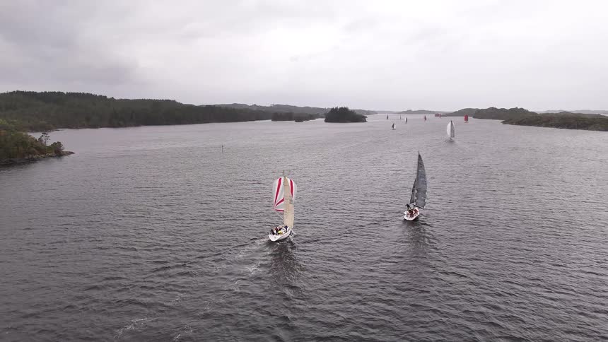 An aerial pullback shot reveals multiple sailboats racing through a scenic Nordic fjord under a cloudy sky