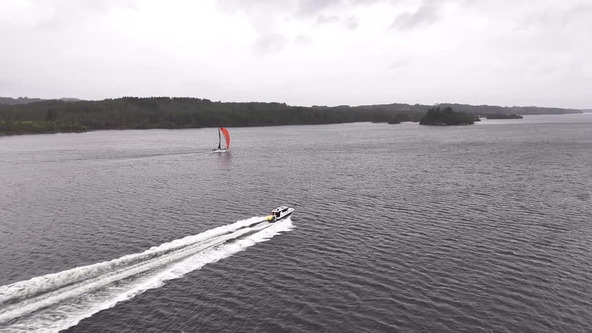 Aerial view of a motorboat speeding across a vast lake towards a sailboat with a bright orange sail on a cloudy day