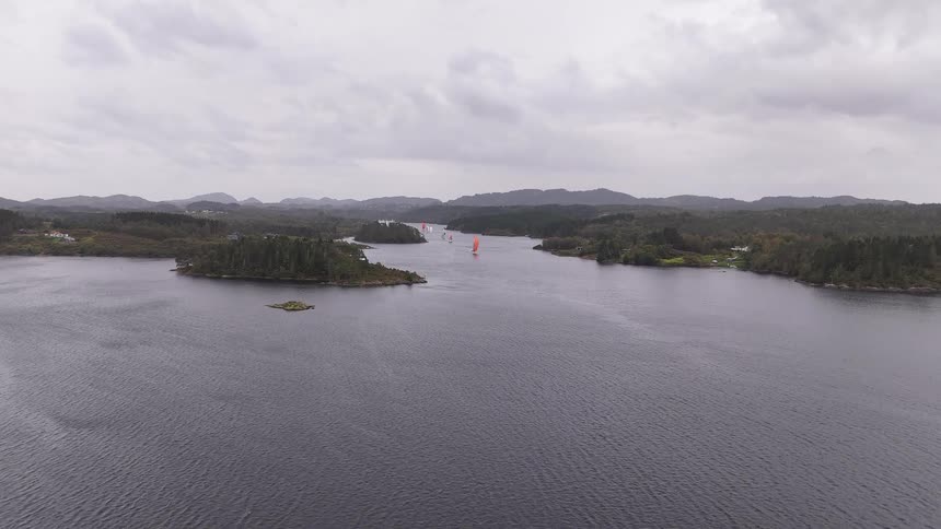 An aerial view slowly advances towards sailboats navigating a vast Norwegian fjord under a cloudy sky