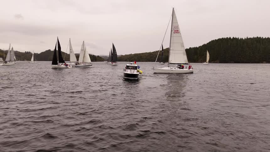 A motorboat cruises among a fleet of sailboats competing in a regatta on a large lake on a cloudy day
