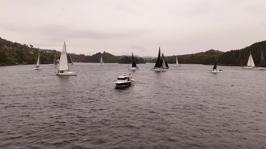 A fleet of sailboats and a motorboat navigate a wide fjord during a regatta on a cloudy day