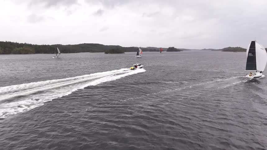An aerial shot follows a speedboat as it cruises past several sailboats during a regatta on a large lake under an overcast sky