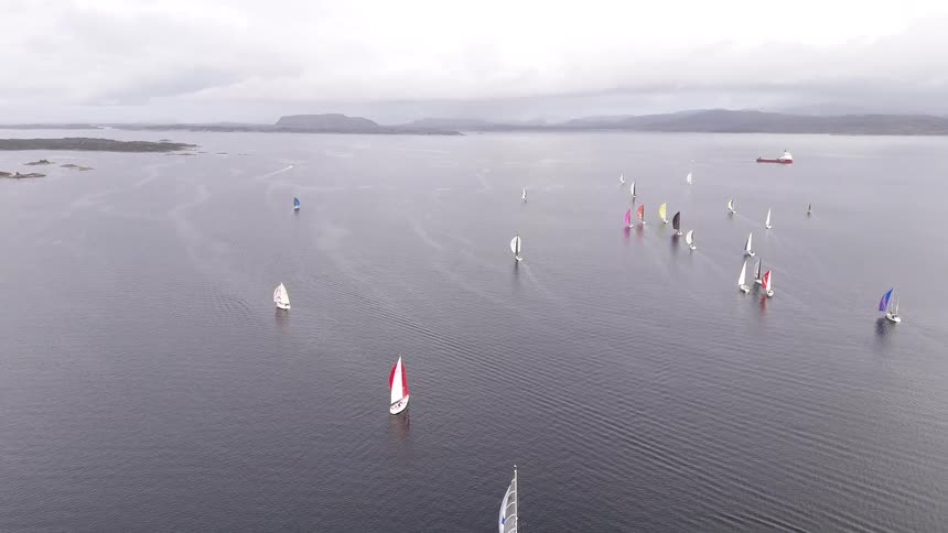 An aerial view shows a fleet of sailboats with colorful spinnakers competing in a regatta on a vast, calm body of water on an overcast day