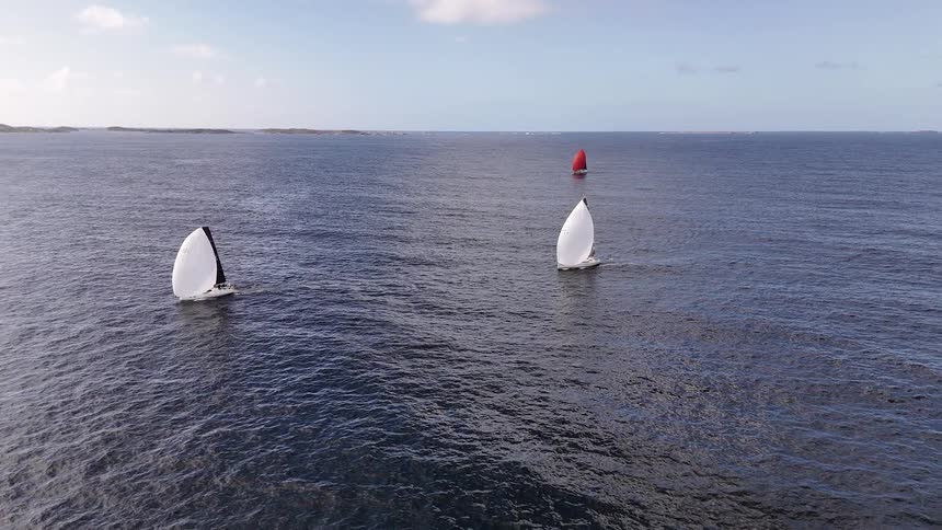 Three sailboats with white and red spinnakers glide across the vast open ocean in a serene aerial shot