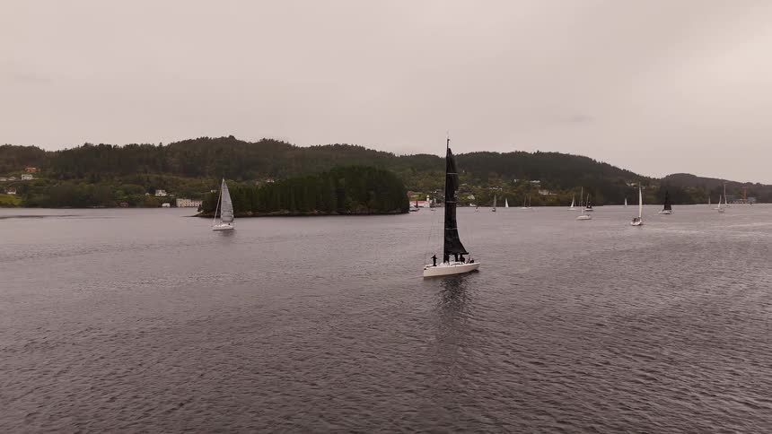 An aerial view shows a fleet of sailboats with black and white sails participating in a regatta on a calm fjord under an overcast sky