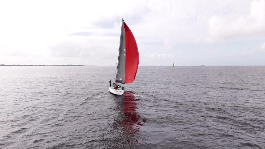 An aerial drone follows behind a sailboat with a striking red spinnaker as it glides across the open ocean on a cloudy day