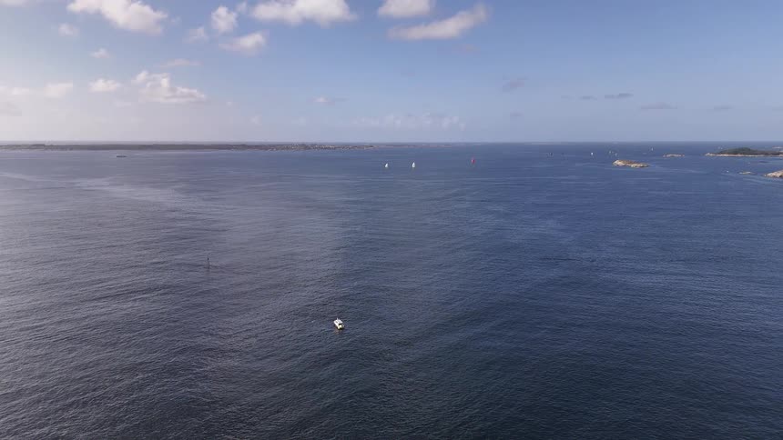 A static, high-angle aerial view shows a vast blue ocean with a few boats and sailboats scattered across the water near a distant archipelago under a partly cloudy sky
