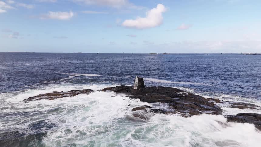 A scenic aerial shot flies slowly towards a stone marker on a rocky islet as waves crash against it in the vast blue ocean