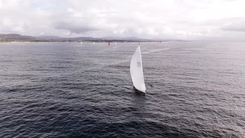 An aerial view captures a sailboat with a large white sail gracefully navigating the open sea during a regatta on a cloudy day