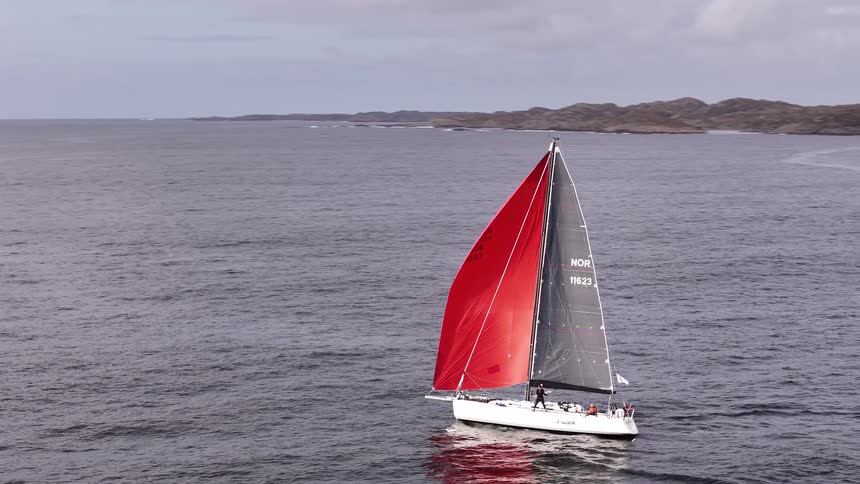 A sailboat with striking red and black sails glides across the open sea towards a rugged, rocky coastline