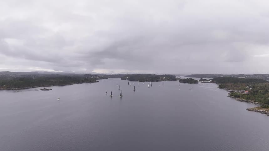 A wide aerial shot captures a sailing regatta taking place in a tranquil fjord surrounded by islands on a moody, overcast day