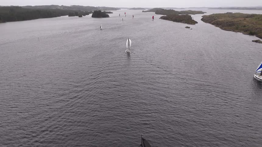 An aerial drone shot follows a fleet of sailboats competing in a regatta on a wide, gray sea on an overcast day