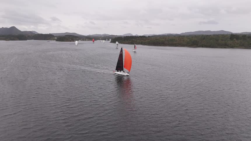 An aerial view follows a sailboat with a vibrant orange and black sail leading a regatta on a cloudy day