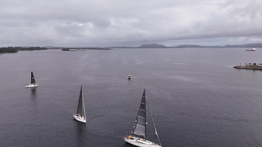 Aerial footage follows a fleet of sailboats racing across a wide, calm fjord on a gray and overcast day