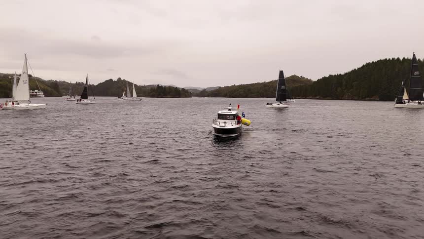 An official motorboat bobs on the water as sailboats compete in a regatta on a vast fjord under an overcast sky